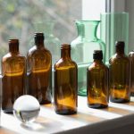 A bright windowsill decorated with various colored glass pieces including amber bottles, green glass vases, and clear glass orbs, all catching sunlight
