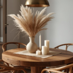 A Homenumental-styled dining corner a round timber table with four rattan chairs, a single dramatic pendant in aged brass, a simple ceramic vase with dried pampas grass, three pillar candles at va