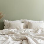 A bedroom wall painted in a soft sage green with warm white linen bedding — demonstrating the calming effect of cool, natural colour on a sleep environment.
