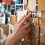 A mood board in progress — hands arranging fabric swatches, paint chips, photographs, and material samples on a cork board. Creative, process-oriented, engaging.
