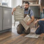 A flooring professional kneeling in a kitchen during an installation consultation, showing material samples including porcelain tile, LVP, and engineered hardwood to a homeowner.